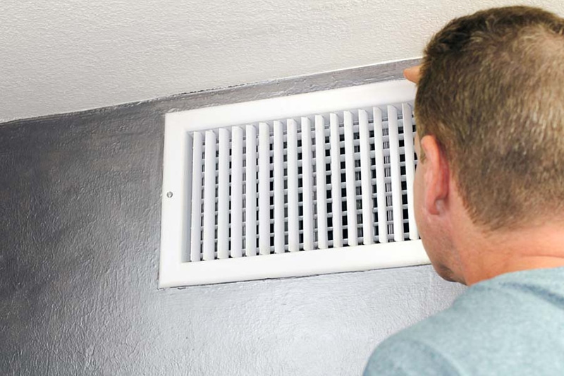Why Is My Air Conditioner Making Noise? Mature male peering inside an upper wall white grid air duct on a silver wall near a white ceiling. A guy inspecting a heating and cooling air register duct for maintenance.