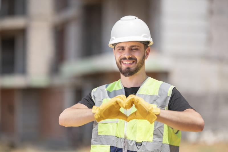 How to Join the HVAC Industry. Image is a photograph of an HVAC professional wearing a white helmet and reflective vest. He is smiling and looking at the camera in front of a construction site. He is making a heart shape with his hands.